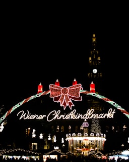 A festive Christmas market scene illuminated at night with a large decorative sign reading 'Wiener Christkindlmarkt'. The sign features a large red bow and several lit candles. Below, a carousel is brightly lit and surrounded by decorated stalls. In the background, a tall clock tower is lit up, with the clock face clearly visible.