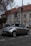 A silver convertible car is parked on a cobblestone street near a building with a red-tiled roof and several windows. A parking sign indicates the end of a parking zone. The street and building have a slightly overcast appearance, with bare tree branches visible in the scene.