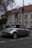 A silver convertible car is parked on a cobblestone street near a building with a red-tiled roof and several windows. A parking sign indicates the end of a parking zone. The street and building have a slightly overcast appearance, with bare tree branches visible in the scene.