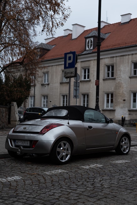 A silver convertible car is parked on a cobblestone street near a building with a red-tiled roof and several windows. A parking sign indicates the end of a parking zone. The street and building have a slightly overcast appearance, with bare tree branches visible in the scene.