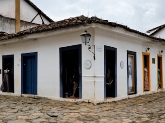 A corner of an old building with a textured stone pathway surrounding it. The walls are painted white with dark blue and orange door frames. Two mannequins in colorful dresses are visible through the doors. A vintage-style street lamp is mounted on the wall above a sign that reads 'N&oacute;s'. The roof is covered with weathered tiles.