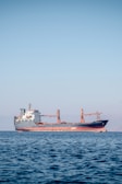 A large cargo ship is floating on a calm sea under a clear blue sky. The ship appears to be carrying containers and is painted red and black, with a white superstructure and cranes on deck. There are distant mountains visible faintly in the background.
