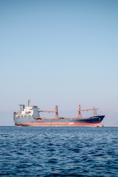 A large cargo ship is floating on a calm sea under a clear blue sky. The ship appears to be carrying containers and is painted red and black, with a white superstructure and cranes on deck. There are distant mountains visible faintly in the background.