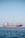A large cargo ship is floating on a calm sea under a clear blue sky. The ship appears to be carrying containers and is painted red and black, with a white superstructure and cranes on deck. There are distant mountains visible faintly in the background.