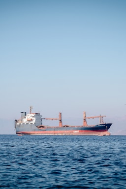 A large cargo ship is floating on a calm sea under a clear blue sky. The ship appears to be carrying containers and is painted red and black, with a white superstructure and cranes on deck. There are distant mountains visible faintly in the background.