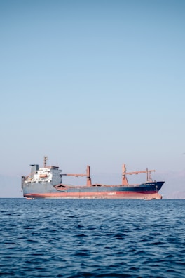 A large cargo ship is floating on a calm sea under a clear blue sky. The ship appears to be carrying containers and is painted red and black, with a white superstructure and cranes on deck. There are distant mountains visible faintly in the background.