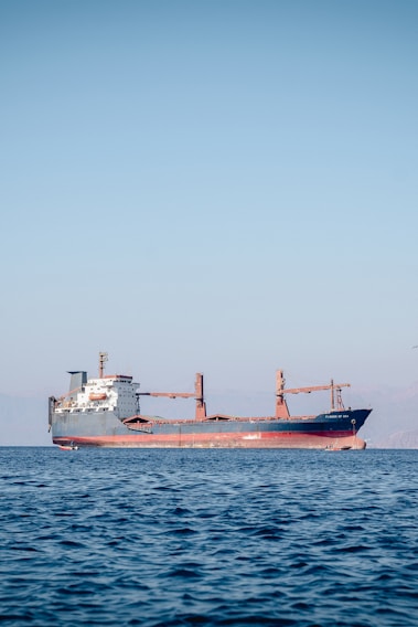 A large cargo ship is floating on a calm sea under a clear blue sky. The ship appears to be carrying containers and is painted red and black, with a white superstructure and cranes on deck. There are distant mountains visible faintly in the background.
