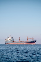 A large cargo ship is floating on a calm sea under a clear blue sky. The ship appears to be carrying containers and is painted red and black, with a white superstructure and cranes on deck. There are distant mountains visible faintly in the background.
