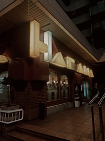 Neon signage with large letters mounted on a dark building exterior. The space includes a brick wall and a sidewalk leading up to a building entrance. Illuminated windows with visible indoor decor and other small, round lights are part of the setup. A potted plant is placed next to the wall, and a metallic railing is present on the entrance path.