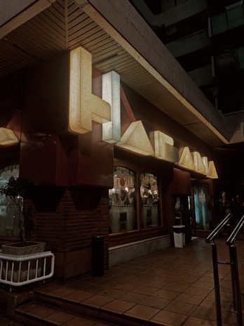 Neon signage with large letters mounted on a dark building exterior. The space includes a brick wall and a sidewalk leading up to a building entrance. Illuminated windows with visible indoor decor and other small, round lights are part of the setup. A potted plant is placed next to the wall, and a metallic railing is present on the entrance path.