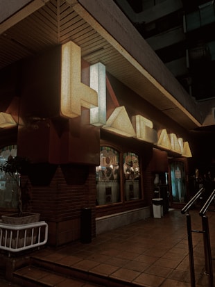 Neon signage with large letters mounted on a dark building exterior. The space includes a brick wall and a sidewalk leading up to a building entrance. Illuminated windows with visible indoor decor and other small, round lights are part of the setup. A potted plant is placed next to the wall, and a metallic railing is present on the entrance path.