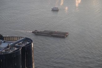 A barge travels across a body of water with a smaller boat in the same vicinity. Nearby, there is the top of a building with visible structures resembling air conditioning units or vents. The water is calm with a few ripples reflecting the light.