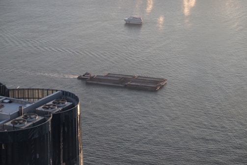 A barge travels across a body of water with a smaller boat in the same vicinity. Nearby, there is the top of a building with visible structures resembling air conditioning units or vents. The water is calm with a few ripples reflecting the light.