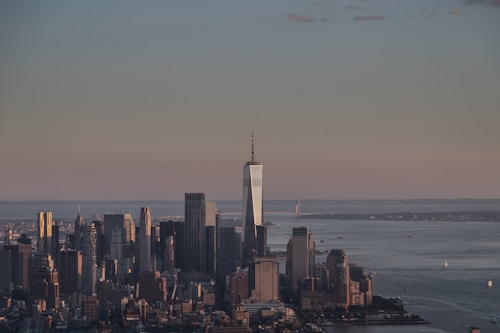 A city skyline of tall, modern skyscrapers under an expansive sky with a calm body of water in the background, reflecting the subdued light of a setting or rising sun.