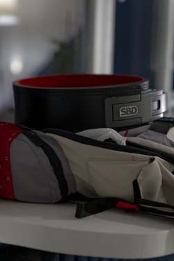 A weightlifting belt with a black exterior and red interior is placed on a surface. Nearby, a gym bag with a mix of gray and red fabric is partially open.