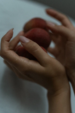 Close-up of a hand holding a peach-colored body lotion with fresh peaches in the background.