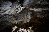 A vast cave with intricate rock formations, including stalactites and stalagmites, extending from the ceiling and floor. The rugged textures of the rocks are illuminated by artificial lighting, creating contrasts and shadows that highlight the natural beauty of the subterranean environment.