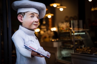 A large, whimsical statue of a chef is positioned outside a bakery. The chef is dressed in a traditional white uniform and hat, holding a box of pastries. The background features a warmly lit interior with shelves displaying various baked goods and soft, yellow lights illuminating the space.