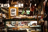 A cozy interior of a delicatessen filled with various hanging cured meats and others displayed on a counter. Framed black and white photographs are adorned on the walls, adding a vintage charm. The display area is surrounded by greenery and other decorative items, creating a warm and inviting atmosphere.