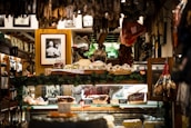 A cozy interior of a delicatessen filled with various hanging cured meats and others displayed on a counter. Framed black and white photographs are adorned on the walls, adding a vintage charm. The display area is surrounded by greenery and other decorative items, creating a warm and inviting atmosphere.