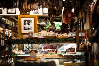 A cozy interior of a delicatessen filled with various hanging cured meats and others displayed on a counter. Framed black and white photographs are adorned on the walls, adding a vintage charm. The display area is surrounded by greenery and other decorative items, creating a warm and inviting atmosphere.