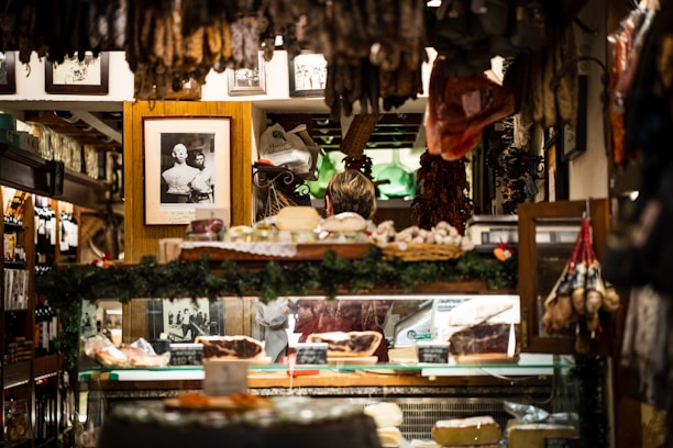 A cozy interior of a delicatessen filled with various hanging cured meats and others displayed on a counter. Framed black and white photographs are adorned on the walls, adding a vintage charm. The display area is surrounded by greenery and other decorative items, creating a warm and inviting atmosphere.