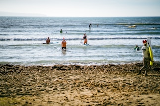 A group of people is enjoying a sunny day at the beach, participating in water sports. Some individuals are standing on paddleboards in the shallow waves, and a few are walking towards the sea with their boards. Everyone is wearing festive red hats, adding a cheerful and holiday-like atmosphere to the scene. The ocean is calm, and the sky is clear, contributing to a pleasant and relaxed mood.