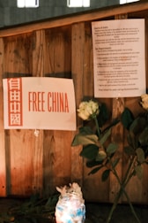 A wooden surface displays a sign with the text 'Free China' in red and white beside a paper listing some requirements or events. In front of the sign, there are white roses and a lit candle in a decorated jar, creating an atmosphere of remembrance or protest.