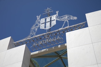 A metal sculpture featuring a kangaroo and an emu holding a shield is positioned above a building with a clear blue sky in the background. The word 'AUSTRALIA' is incorporated into the structure beneath the animals.