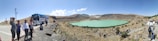 A group of people stand on a rocky path near a tour bus, overlooking a turquoise lake set in a barren, hilly landscape. The sky is clear and blue, and the terrain is mostly dry with sparse vegetation.