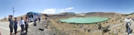 A group of people stand on a rocky path near a tour bus, overlooking a turquoise lake set in a barren, hilly landscape. The sky is clear and blue, and the terrain is mostly dry with sparse vegetation.