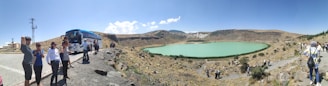 A group of people stand on a rocky path near a tour bus, overlooking a turquoise lake set in a barren, hilly landscape. The sky is clear and blue, and the terrain is mostly dry with sparse vegetation.