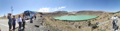A group of people stand on a rocky path near a tour bus, overlooking a turquoise lake set in a barren, hilly landscape. The sky is clear and blue, and the terrain is mostly dry with sparse vegetation.