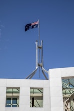 A flag with the British Union Jack and Southern Cross constellation flutters atop a tall metal pole on an architectural structure with white marble tiles against a clear blue sky.