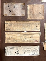 A collection of vintage comic strips from newspapers, including 'Peanuts,' 'Garfield,' and others featuring humorous dialogue and illustrations. The comics are taped onto a brown background, presenting various scenarios involving children, pets, and family life.