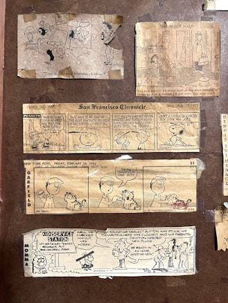 A collection of vintage comic strips from newspapers, including 'Peanuts,' 'Garfield,' and others featuring humorous dialogue and illustrations. The comics are taped onto a brown background, presenting various scenarios involving children, pets, and family life.