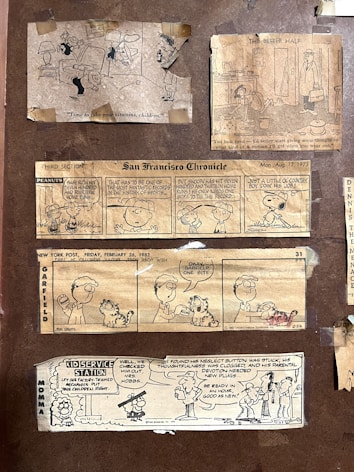 A collection of vintage comic strips from newspapers, including 'Peanuts,' 'Garfield,' and others featuring humorous dialogue and illustrations. The comics are taped onto a brown background, presenting various scenarios involving children, pets, and family life.