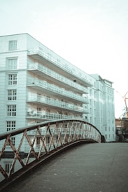 A modern, multi-story apartment building with white facades and numerous balconies is juxtaposed with an old-fashioned, rusted metal bridge in the foreground. The sky above is clear, and there is a construction crane visible in the background, suggesting urban development.