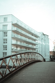 A modern, multi-story apartment building with white facades and numerous balconies is juxtaposed with an old-fashioned, rusted metal bridge in the foreground. The sky above is clear, and there is a construction crane visible in the background, suggesting urban development.