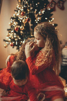 A warm scene with children wearing red dresses, sitting together near a decorated Christmas tree adorned with lights and ornaments. The soft lighting and tender embrace of the children create a cozy, festive atmosphere.