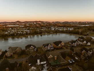 A residential neighborhood with houses lining a large pond. The scene is set against a backdrop of rolling hills under a soft twilight sky. The houses are spacious, with varied architectural styles and well-kept lawns. The pond reflects the warm colors of the setting sun, adding a tranquil ambiance to the landscape.