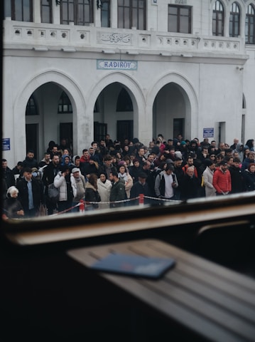 A large group of people gathered in front of a building with arched doorways and windows. The crowd appears diverse, with individuals wearing various types of clothing, including winter jackets. The building has a sign that reads 'Kadikoy' above the entrance.
