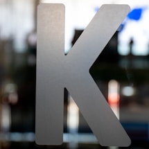 A large, metallic letter 'K' is prominently displayed against a blurred background. The background appears to be a combination of abstract shapes and colors, giving a sense of depth and movement.