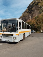 A white bus with a yellow stripe is parked on a paved area. The destination sign reads 'Funchal'. Behind the bus, there are steep, rocky hills covered with brown and green vegetation. A few bare trees are visible against a blue sky with scattered clouds. A small car is parked to the right.