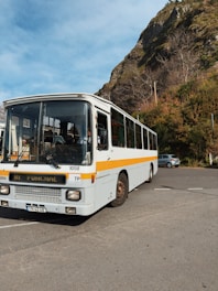 A white bus with a yellow stripe is parked on a paved area. The destination sign reads 'Funchal'. Behind the bus, there are steep, rocky hills covered with brown and green vegetation. A few bare trees are visible against a blue sky with scattered clouds. A small car is parked to the right.