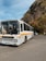 A white bus with a yellow stripe is parked on a paved area. The destination sign reads 'Funchal'. Behind the bus, there are steep, rocky hills covered with brown and green vegetation. A few bare trees are visible against a blue sky with scattered clouds. A small car is parked to the right.