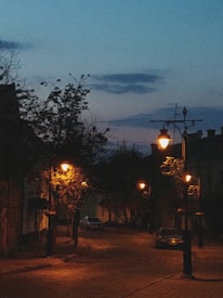 A quiet, dimly lit street with old-fashioned street lamps casting a warm, orange glow on the cobblestone pavement. Sparse trees line the street, and a few parked cars are visible in the background. The sky is transitioning from day to night, with visible shades of blue and gentle clouds.