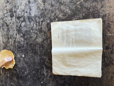 A crumpled book page lies open on a textured, dark surface. The page appears to be from a vintage book with black printed text, slightly yellowed with age. Nearby, a wooden tool or handle extends from a light, circular object resembling a small piece of dough or clay.