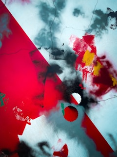 An abstract artwork featuring bold splashes of red, black, and white with hints of yellow and green. The composition includes sharp angles and blurred edges, with various overlapping textures that create a dynamic and chaotic visual effect.