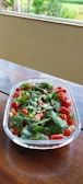 A clear glass bowl filled with a fresh salad consisting of cherry tomatoes, basil leaves, and pieces of cheese sits on a wooden table. The vivid colors of the ingredients stand out against the backdrop of a green garden visible through a large window.